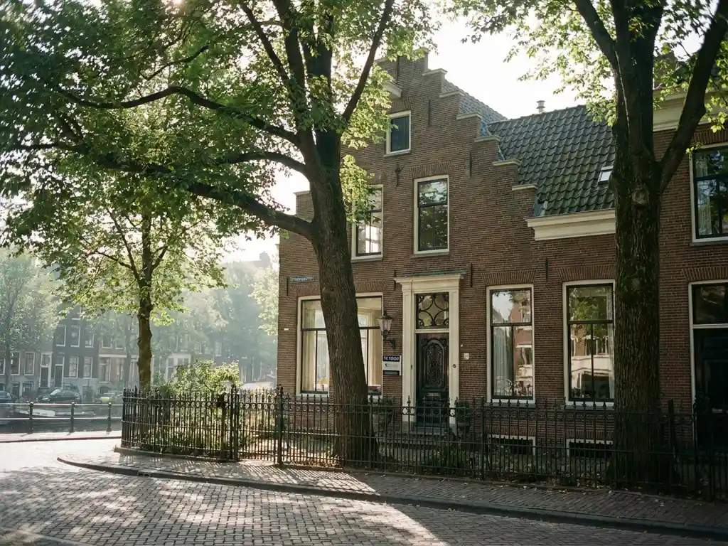 Nederlands grachtenpand met trapgevel achter bomen, "Te Koop" bordje bij voordeur, ochtendlicht op kinderkopjes