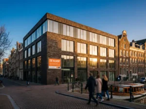 Modern kantoorgebouw met glazen gevel en bakstenen facade toont "Te Koop" bord aan Amsterdamse gracht in warme middagzon