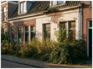 Vervallen Nederlands rijtjeshuis met afbladderende witte verf, gebarsten bakstenen gevel en overwoekerde tuin tijdens gouden uur