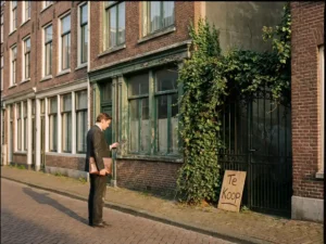 Professionele koper in donker pak onderzoekt verweerd bakstenen herenhuis met Te Koop bord en overwoekerde tuin