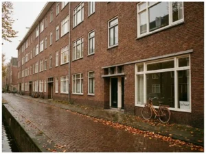 Amsterdams huurappartement in bakstenen jaren 50-stijl aan grachtenstraat, met "te huur"-bordje in raam en herfstbladeren op kinderkopjes.