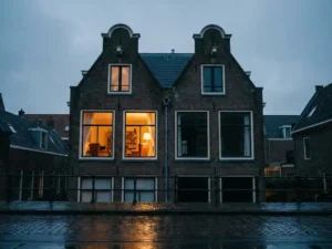 Amsterdams grachtenpand met verlichte woonkamer naast leegstaande donkere eenheid, weerspiegeld in natte kinderkopjes.