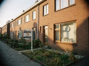 Nederlandse rijtjeshuis met Te Koop-bord, krimpende eurobiljetten naast een klok op vensterbank, rode bakstenen gevel in middaglicht.
